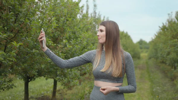 Joyful fitness woman during live stream broadcasting. A modern sports woman takes pictures on the nature smartphone, recording a video blog. Sports video vlog. Video Blog athlete. Girl blogger