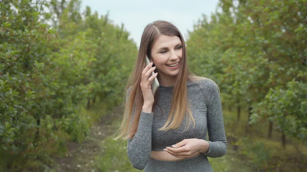 Onnellinen hipster nainen kirjoittaa matkapuhelimella ulkona. Iloinen tyttö älypuhelin puistossa taustalla puita. Hymyilevä nainen pitelee kännykkää ulkona. Tyttö kommunikoi puhelimitse — kuvapankkivalokuva