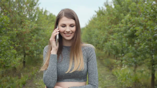Onnellinen hipster nainen kirjoittaa matkapuhelimella ulkona. Iloinen tyttö älypuhelin puistossa taustalla puita. Hymyilevä nainen pitelee kännykkää ulkona. Tyttö kommunikoi puhelimitse — kuvapankkivalokuva
