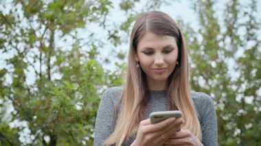 Dışarıda cep telefonuyla yazan mutlu kadın. Arka planda akıllı telefonu olan neşeli bir kız. Dışarıda cep telefonunu tutan gülümseyen kadın. Spor kızı, internet teknolojisi, koşucu egzersizi.