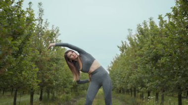 Kadın koşucu, yaz parkında egzersiz yapmadan önce kollarını esnetiyor. Orta yaşlı, atletik bir kadın, koşuya çıkmadan önce vücudu ısıtıyor. Beyaz şortunu giyip koşuyor.