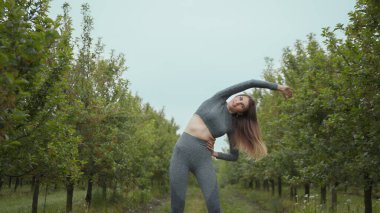 Kadın koşucu, yaz parkında egzersiz yapmadan önce kollarını esnetiyor. Orta yaşlı, atletik bir kadın, koşuya çıkmadan önce vücudu ısıtıyor. Beyaz şortunu giyip koşuyor.