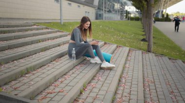 Fitness Kadını telefonda egzersiz uygulaması kullanıyor. Merdivenlerde oturarak egzersiz yapıyor. Spor ve Teknoloji, Kilo Verme ve Eğitim. Spor salonunda dinlenirken cep telefonu kullanan kadın