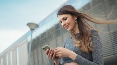 Spor salonundaki genç ve güzel bir kızın elinde akıllı bir telefon tutarak spor uygulaması kullanmasının yakın çekimi. Modern Şehir Sokakta Çekim. Açık havada koşarken akıllı telefona bakan portre kadını.