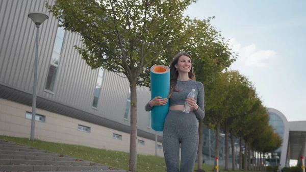 Active woman with fitness mat and water bottle in hands walking in park. Happy lady strolling in summer city park going at outdoors yoga class. Active people. Fitness girl. Inspiration and motivation