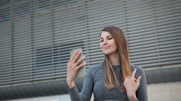 Nær den unge, hvite kvinnen som har en videochat om den moderne bygningens bakgrunn. Utendørs. POV til den sjarmerende og lykkelige jenta. Nærbilde av en vakker sportsjente som holder en smarttelefon – stockfoto