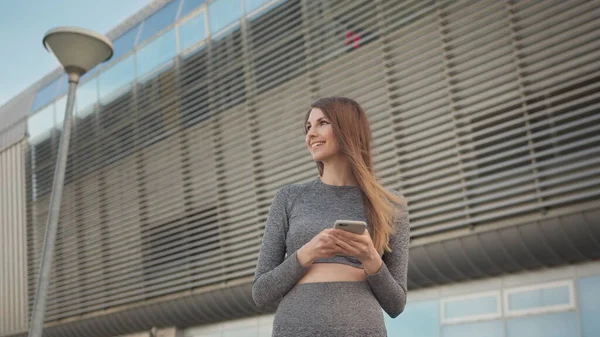 Portrettdame ser på smarttelefon mens hun løper utendørs. Nær-up av en ung, vakker sportsjente som holder en smarttelefon i hendene og bruker en støtfangerassistent. Modern City Shooting On the Street – stockfoto