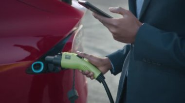 Takım elbiseli Afrikalı adam modern elektrikli arabayı şarj ederken akıllı telefondan acil sorunları çözüyor. İşadamı dışarıda lüks kırmızı arabasının yanında duruyor. Akıllı telefon kullanan adam elektrikli araçlara bağlanıyor.