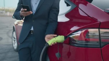 Başarılı iş adamı istasyonda elektrikli araba şarj ediyor ve modern akıllı telefonu iş için kullanıyor. Takım elbiseli Afrikalı adam dışarıda duruyor, gülümsüyor ve telefona bakıyor. Elektrikli arabaya akıllı telefon bağlantısı