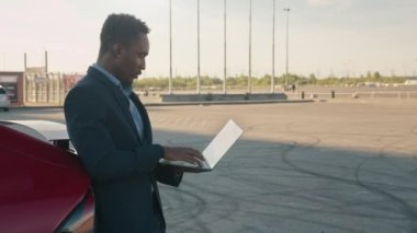 Yaklaş, Afro-Amerikan adam elinde kablosuz laptop tutarken lüks kırmızı arabasına yaslanıyor. İş adamı arkadaşıyla Skype 'ta konuşuyor. Kendi lüks elektromobil durak şarj istasyonu, şarj istasyonu...