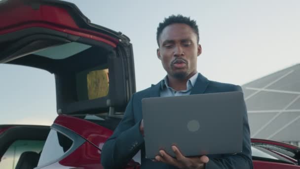 En afroamerikan som håller i trådlös laptop medan han lutar sig mot sin lyxiga röda bil. Ung man i kostym väntar medan elbil förnya energi i batteri. Man talar via video på bärbar dator — Stockvideo