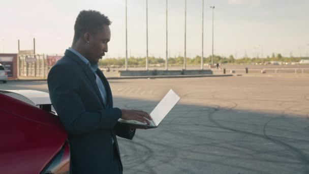 Da vicino, un uomo afroamericano con un portatile senza fili mentre si appoggia alla sua auto rossa di lusso. Uomo d'affari che parla su skype con un amico quando proprio caricabatterie elettromobile di lusso, stazione di ricarica — Video Stock