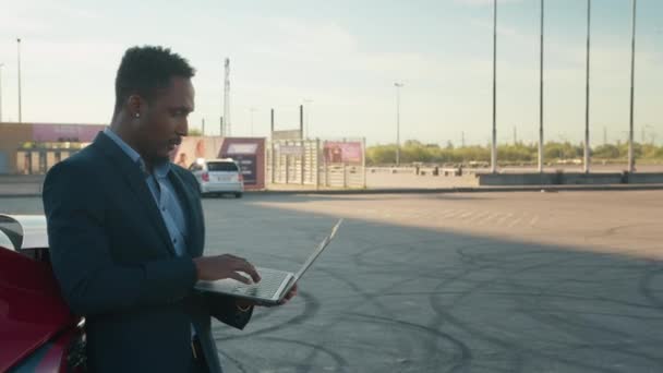 Uomo d'affari che parla su skype con un amico quando proprio caricabatterie elettromobile di lusso, stazione di ricarica. Da vicino, l'uomo afro-americano con in mano un portatile senza fili mentre si appoggia alla sua auto rossa di lusso — Video Stock