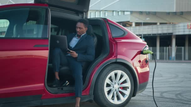 高級電気自動車が充電されている間、リモートワークのための現代のラップトップを使用してスタイリッシュなスーツのアフリカ系アメリカ人ビジネスマン。ハンサムな男性座っていますサイドオートでオープンドアと探してラップトップ. — ストック動画
