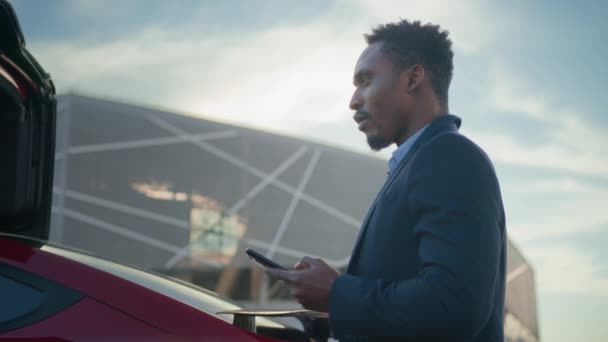 一位身穿西服的非洲裔美国商人站在户外用电动汽车收费电缆使用现代智能手机进行了近距离采访。现代技术、商业和人的概念. — 图库视频影像