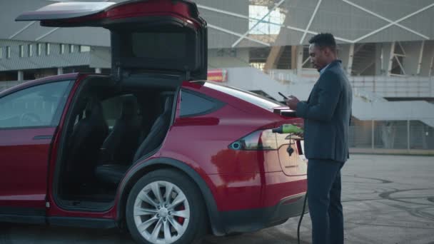 Hombre africano serio sosteniendo cable de carga en la mano y teléfono inteligente moderno en otro, mientras que de pie cerca de coche eléctrico de lujo. Joven hombre en traje de negocios posando al aire libre y mirando smartphone. — Vídeos de Stock