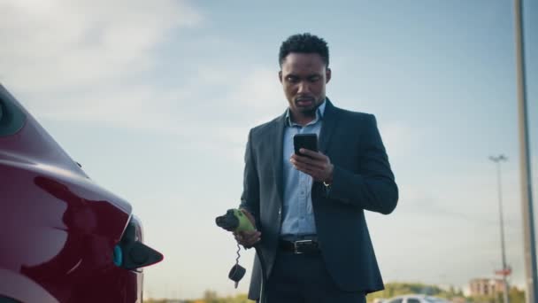 Primo piano di uomo d'affari africano scollegare cavo di ricarica da auto elettrica durante l'utilizzo di smartphone moderno. Concetto di stili di vita, veicolo ecologia e persone occupate. Collegamento del cavo di alimentazione a un'auto. — Video Stock