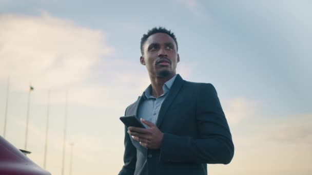 Giovane uomo d'affari di successo ricarica auto elettrica sulla stazione e utilizzando smartphone moderno per il lavoro. Uomo africano in giacca e cravatta in piedi all'aperto, sorridente e guardando altrove. — Video Stock