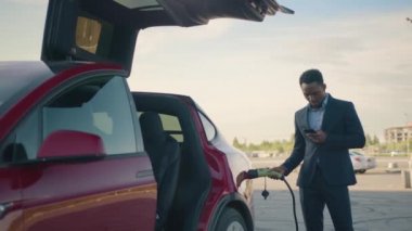 Sıradan bir plan, takım elbiseli yakışıklı bir Afrikalı 'nın portresi bavulu tutuyor ve dışarıda elektrikli araba şarj ediyor. İnsan kavramı, modern yaşam tarzları ve çevre dostu araç. Şarj edilen araba