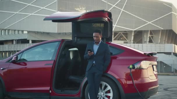 Красивий чоловік використовує мобільний телефон під час заряджання батареї свого червоного електромобіля. Впевнений африканський чоловік у діловому костюмі стоїть біля розкішного автомобіля з сучасним смартфоном в руках. Зарядний автомобіль — стокове відео