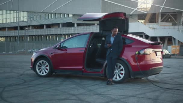 Un Africain attend pendant que sa voiture électrique se charge. Je charge la voiture. Portrait complet de l'homme d'affaires réussi en costume en utilisant un smartphone moderne pour vérifier les e-mails de travail tout en s'appuyant sur la voiture rouge. — Video