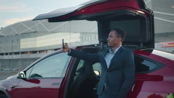 Empresario afroamericano sonriendo joven hombre de negocios haciendo videollamada selfie de pie al aire libre en el distrito de negocios. Retrato de un bloguero de negocios en la estación de carga del coche eléctrico — Vídeos de Stock