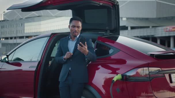 African man, standing near luxury charging electric car and talking on mobile. Male showing sign OK with fingers and smiling on smartphone. Male entrepreneur having working meeting through video call — Stock Video