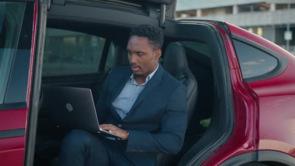 ノートパソコンの男が電気自動車の充電中に座っている。赤い車の中に座っている男性起業家は、ドアを開けて膝の上にポータブルノートパソコンを折り畳みます。プラグに充電ケーブル付き電気自動車. — ストック動画