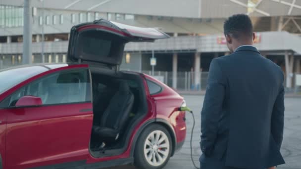 Afrikaanse zakenman die luxe elektrische auto oplaadt. Succesvolle Afrikaanse zakenman met koffer terwijl past in de buurt van elektrische auto met oplaadkabel in plug. Milieuvriendelijk opladen van voertuigen op het station — Stockvideo