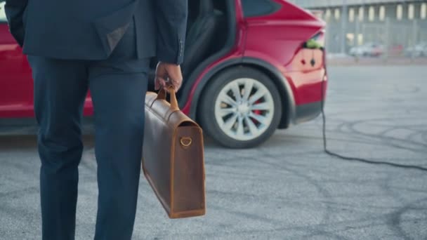 Gros plan de l'homme afro-américain en costume formel reliant le câble de charge à la voiture électrique. Jeune homme debout près de sa voiture moderne avec valise en cuir à la main. Connexion de charge à la voiture électrique. — Video