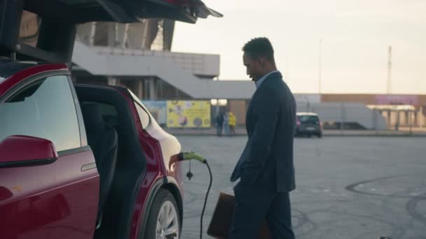 Hombre de negocios africano cargando coche eléctrico de lujo. Exitoso empresario africano sosteniendo la maleta mientras se adapta cerca del coche eléctrico con cable de carga en el enchufe. Carga de vehículos ecológicos en la estación — Vídeos de Stock