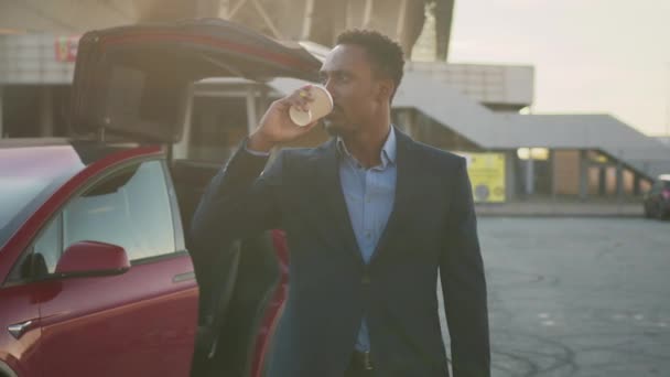 Portret odnoszącego sukcesy afrykańskiego biznesmena w stylowym garniturze pijącego kawę, podczas gdy jego luksusowy czerwony samochód elektryczny się ładuje. Szczęśliwy mężczyzna uśmiechający się i patrzący w kamerę. Biznesmen samochód elektryczny ładuje — Wideo stockowe