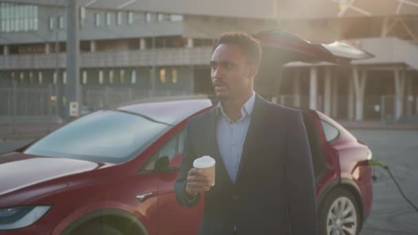 Hombre africano bebiendo café mientras el coche eléctrico se carga. Hombre africano guapo en traje de negocios bebiendo café fresco mientras espera a que se cargue el coche eléctrico moderno. Carga automática eléctrica. — Vídeos de Stock