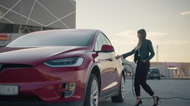 Joven hermosa Chica con estilo se acerca a un lujoso coche eléctrico moderno, que se carga en la estación de carga. La chica abre la puerta automática del coche en el botón y se mete en el coche — Vídeo de stock