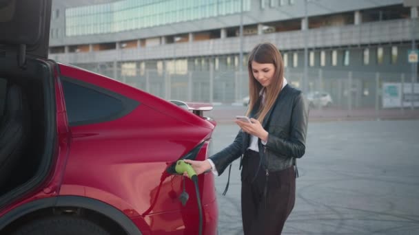 Mulher bonita usando smartphone enquanto carrega carro. Mulher nova no desgaste casual que paga em linha para carregar o veículo. Mulher branca usando smartphone moderno e carregando carro elétrico ao ar livre — Vídeo de Stock