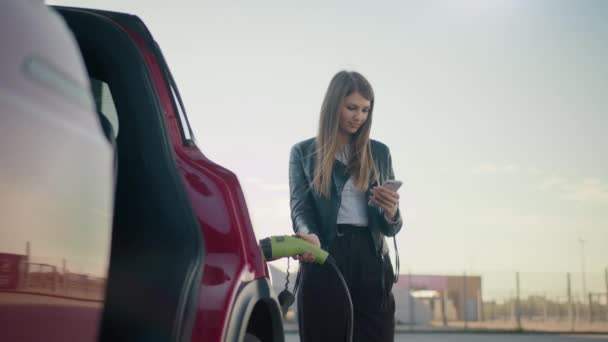 漂亮的高加索女人使用现代智能手机，在户外为电动车充电。穿着休闲装的年轻女性在网上支付车费。充电时使用智能手机的女人 — 图库视频影像