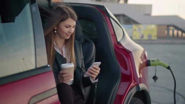 Retrato Mulher encantadora sentada dentro do carro elétrico de carregamento com smartphone moderno e xícara de café nas mãos. Jovem menina moderna olha do telefone para a câmera e sorri. À espera de carro para cobrar — Vídeo de Stock