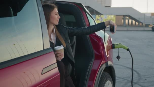 Γοητευτική γυναίκα κάθεται μέσα φόρτιση ηλεκτρικό αυτοκίνητο με σύγχρονο smartphone και φλιτζάνι καφέ στα χέρια. Λευκή γυναίκα που μιλάει μέσω βιντεοκλήσης ή βγάζει selfie σε smartphone. Γυναίκα που κάθεται στο αυτοκίνητο — Αρχείο Βίντεο