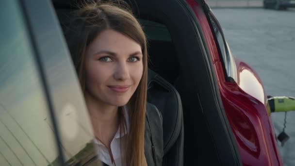 Πορτρέτο Γοητευτική γυναίκα κάθεται μέσα φορτίζοντας ηλεκτρικό αυτοκίνητο. Νεαρή μοντέρνα κοπέλα κοιτάζει από το τηλέφωνο στην κάμερα και χαμογελά. Πορτρέτο της χαρούμενης νεαρό κορίτσι γέλιο, ενώ κάθεται στο αυτοκίνητο — Αρχείο Βίντεο