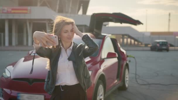 Beyaz bir kadın lüks bir kırmızı arabanın önünde akıllı bir telefondan selfie çekiyor. Elektrikli arabayı şarj eden ve akıllı telefon kullanan bir kadın internetteki bloglarda fotoğraflanıyor. Çevre dostu araç kavramı — Stok video
