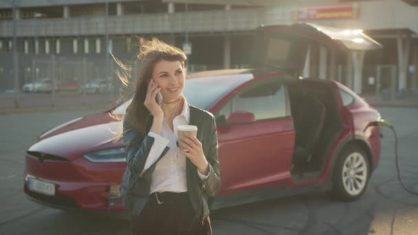 Mulher ter chamada no smartphone enquanto carrega carro. Mulher caucasiana encantadora que tem chamada no smartphone moderno enquanto carrega seu carro elétrico de luxo. Conceito de gadgets de pessoas e veículo ecológico — Vídeo de Stock