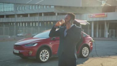 Modern elektrikli arabası şarj olurken Afrikalı Amerikalı işadamı cep telefonuyla konuşuyor ve kahve içiyor. Kendine güvenen şık takım elbiseli genç adam beklerken mesafeli çalışıyor. Elektrikli araba.