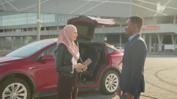 Uomo e donna musulmana in hijab in piedi insieme all'aperto vicino auto elettrica di lusso. Acquisto di veicoli ecologici. Uomo d'affari africano di successo in tuta esaminando veicolo costoso prima dell'acquisto. — Video Stock