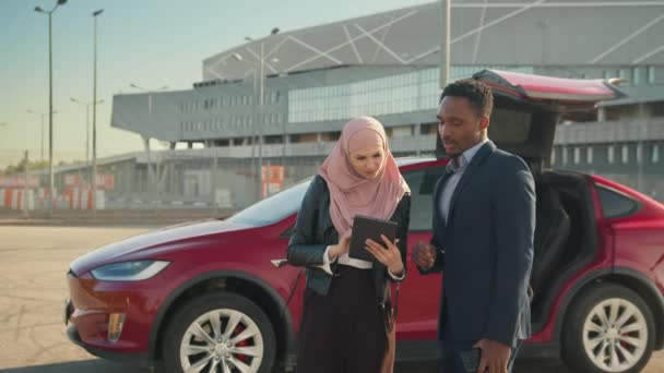 Mujer musulmana en hiyab usando tableta digital mientras habla con un hombre de negocios africano de traje. Mujer vendedor y cliente masculino de pie cerca de coche eléctrico y discutir todos los detalles de la compra costosa. — Vídeos de Stock