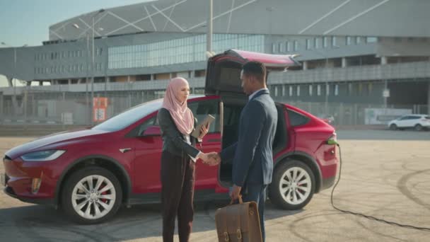 Donna musulmana in hijab che vende auto elettriche di lusso a uomo d'affari africano in suit.Competent rivenditore di auto femminili in possesso di tablet digitale e stringendo la mano con il cliente maschile. Donna che vende auto elettriche — Video Stock