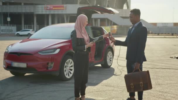 Мусульманская женщина дает ключи от электромобиля африканскому мужчине. Улыбающаяся мусульманка в хиджабе дает ключи от электромобиля африканскому бизнесмену в костюме. Счастливый клиент покупает экологически чистый автомобиль — стоковое видео