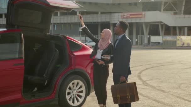 Distribuidor femenino musulmán mostrando coche eléctrico rojo al cliente africano al aire libre. Hombre africano en traje de negocios comprando vehículo ecológico. Concepto de venta y compra. — Vídeos de Stock