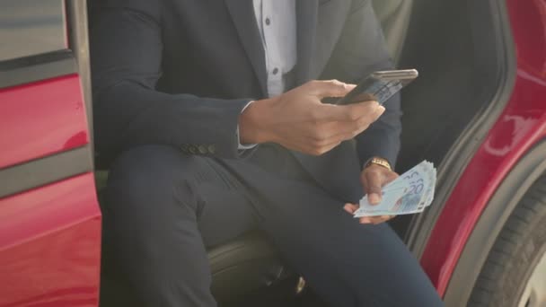 Mannen sitter i elbil med mobil och pengar kontanter. Allvarlig afrikansk affärsman sitter i bil med öppen dörr och håller smartphone med pengar kontanter. Lyxbilen laddar. Begreppet besparingar — Stockvideo