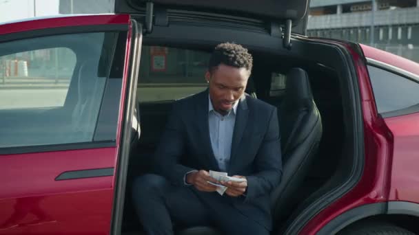 充電している電気自動車の中に座って手に現金でビジネススーツのハンサムなアフリカ人男性。人、輸送、貯蓄の概念。お金のある男電気自動車に座って — ストック動画