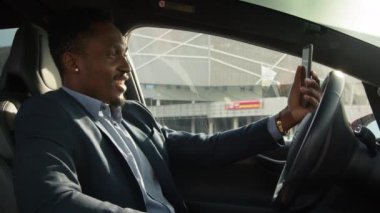 Takım elbiseli yakışıklı Afrikalı bir adam lüks arabasının şoför koltuğunda oturuyor, kahve içiyor ve modern akıllı telefon kullanıyor. Genç bir erkek cep telefonundan iş e-postalarını kontrol ediyor. Video çağrısı